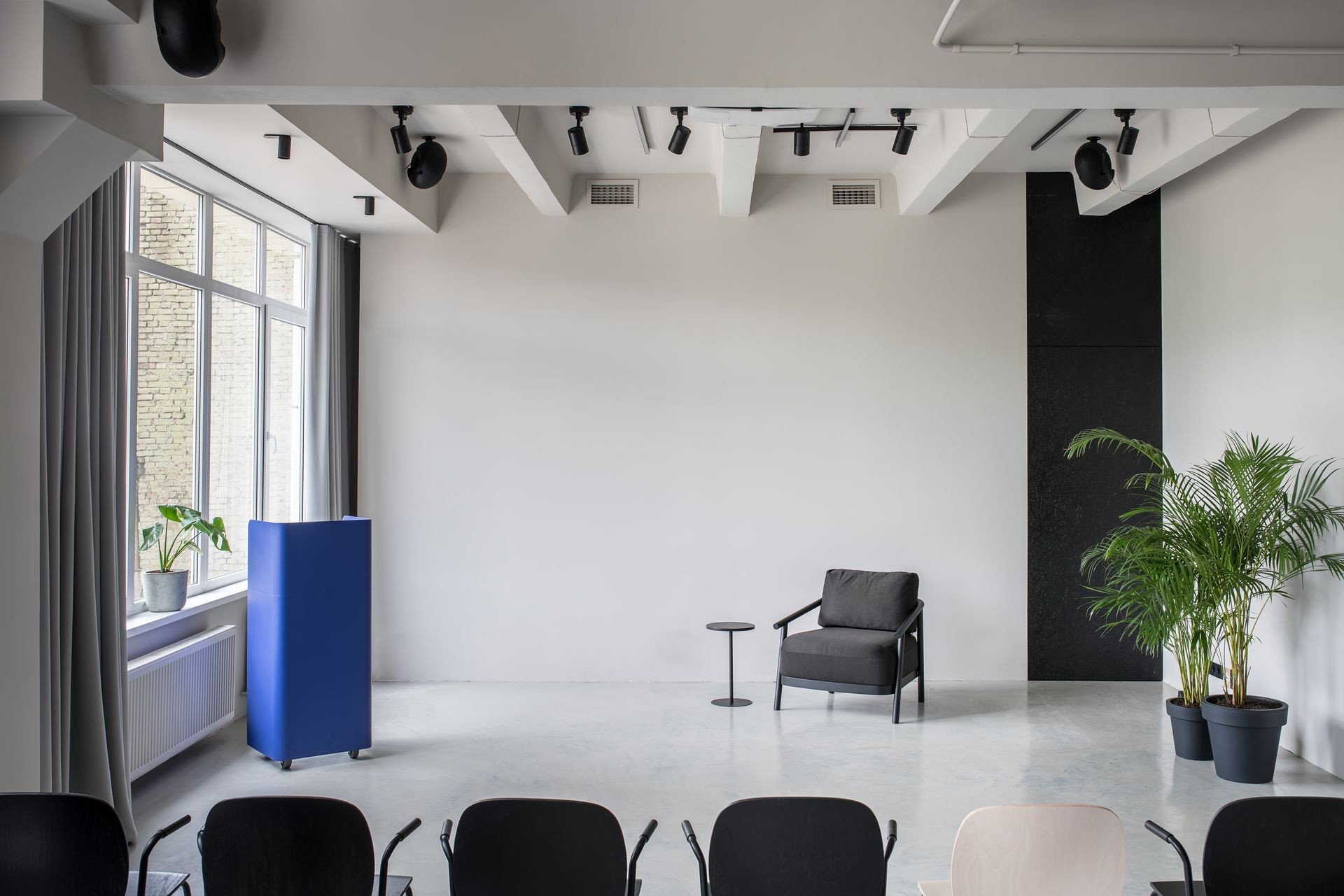 Trendy conference hall with gray walls and a glossy floor. There is a row of multicolor chairs, metallic blue stand,dark armchair, small round table, green plants in the pots, window with curtains.