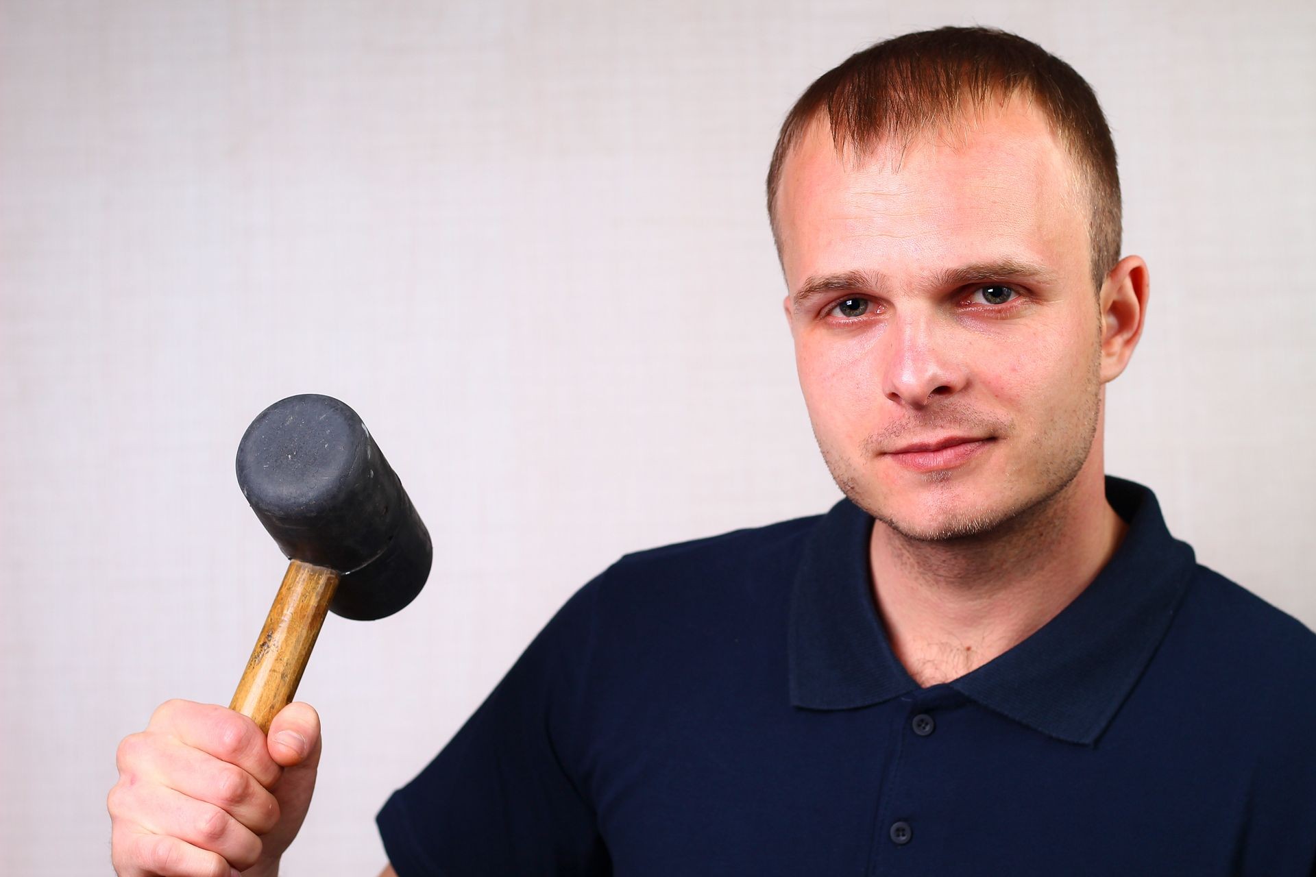 A working professional finisher in a classic blue t-shirt with a collar holds a rubber mallet in his hand. A confident look, a symbol of warranty and reliability.